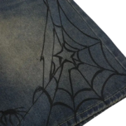 Close-up of Sp5der brand denim shorts featuring a black printed spider web graphic detail on blue distressed denim fabric.