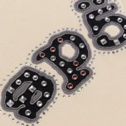 Close-up of the black Sp5der logo appliqué with metal studs and spikes on a cream Mixed Stone OG V2 zip hoodie.