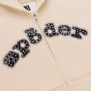 Close-up view of the Sp5der Mixed Stone OG V2 Cream zip hoodie showcasing the logo with metal studs and crystal details.