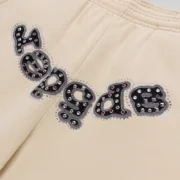 Close-up of Sp5der Mixed Stone OG V2 Cream sweatpants featuring an embellished logo appliqué with metal studs.