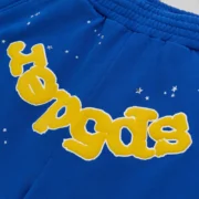 Close-up view of the Sp5der Blue OG Logo Sweatshort featuring yellow text print and white star graphics on cotton fleece.
