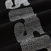 Close-up of the sparkling rhinestone logo detail on the black Sp5der silk sweatpants apparel product texture.