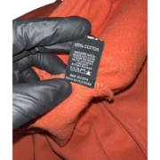 Hand in a black glove holding the interior care tag of an orange Sp5der Worldwide sweat short, 100% cotton material.