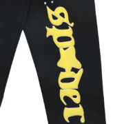 Close-up view of the Sp5der Jeffery sweatpants featuring a large vertical yellow logo print on black fabric.