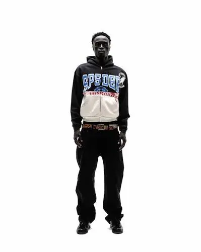 Model wearing a black and white Sp5der zip-up hoodie with blue lettering, black pants, and a patterned belt