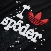 Detailed close-up of the Sp5der x Adidas Souvenir Tee in black featuring a bold white graphic print and red Trefoil logo.