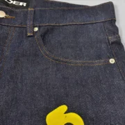 Close-up view of the premium Sp5der Beluga Jean in dark indigo denim fabric with visible yellow graphic detailing.