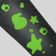 Close-up view of Sp5der Beluga Stamp Denim in Black featuring vibrant neon green star and logo graphic print details.