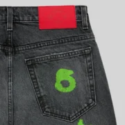 Close-up of Sp5der Beluga Stamp Denim in faded black, featuring a neon green stamp graphic and red branded leather patch.