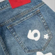 Close-up view of the back pocket on Sp5der Beluga Wash Denim in Light Indigo with a white V3 graphic and red brand tag.