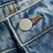 Close-up view of the branded metal button on Sp5der Beluga Wash Denim in Light Indigo, featuring detailed stitching.