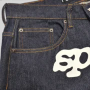Close-up view of the Sp5der V3 Indigo Denim jeans showcasing the premium fabric texture and front graphic detail.