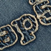 Close-up view of the frayed embroidered SP5DER logo on the Sp5der Denim Work Jacket in Light Blue Wash finish.