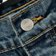 Detail shot of the custom branded metal button on Sp5der Light Blue Wash denim work pants with contrast stitching.