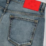 Close-up of Sp5der Light Blue Wash denim work pant featuring the signature red branded leather back patch detail.