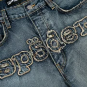 Close up of the Sp5der Denim Work Pant in Light Blue Wash showing the signature frayed embroidered logo detailing.
