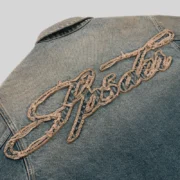 Close-up of the light wash Sp5der Script Denim Work Jacket featuring signature frayed embroidered script logo detailing.