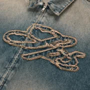 Close-up of the light wash denim Sp5der Script Work Jacket featuring frayed embroidered logo detailing and zip closure.