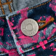Close-up view of the custom branded SP5DER metal button on the Indigo V2 Web Denim jeans with all-over print.