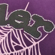 Detailed close-up of the Sp5der Purple Web Hoodie showing black logo text and pink web graphics with rhinestones.